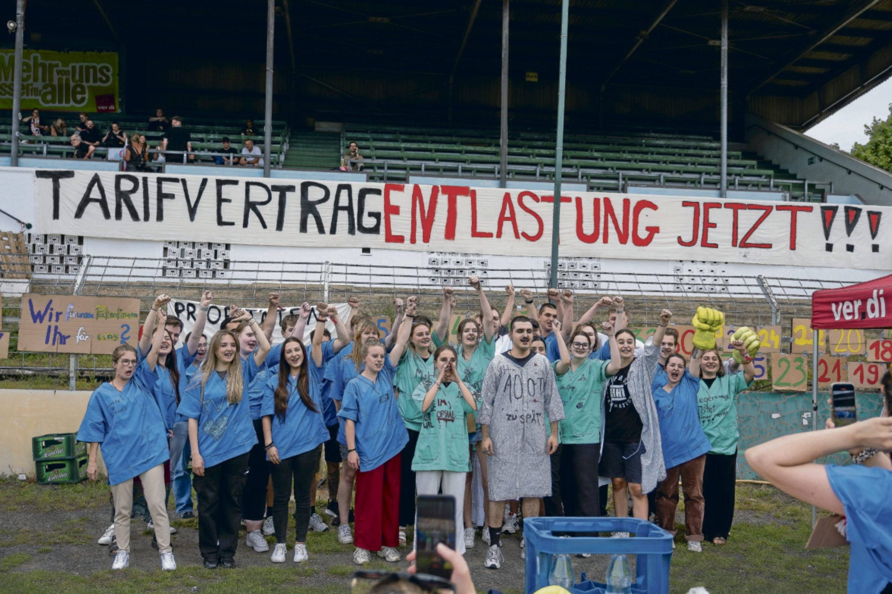 Tarifstreik an Klinikum, Im Hintergrund Plakat mit der Aufschrift 'Tarifvertrag: Entlastung Jetzt!!, Im Vordergrund Menschen in Krankenhausbekleidung die Hände in die Luft streckend